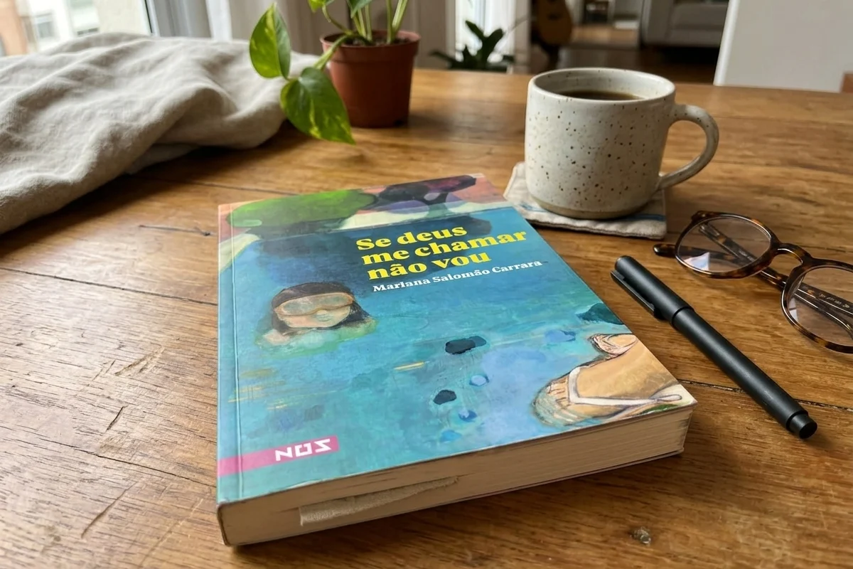 Livro “Se deus me chamar não vou”, de Mariana Salomão Carrara, aberto sobre uma mesa de madeira rústica de um café, ao lado de um latte, óculos e um caderno de notas aberto com caneta.