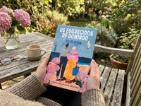 Pessoa a segurar o livro “Os Esquecidos de Domingo”, de Valérie Perrin, num jardim, com uma xícara de café, óculos e hortênsias cor-de-rosa sobre uma mesa de madeira ao fundo.