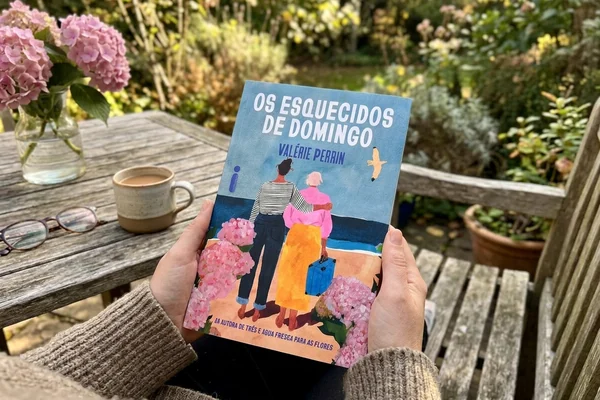 Pessoa a segurar o livro “Os Esquecidos de Domingo”, de Valérie Perrin, num jardim, com uma xícara de café, óculos e hortênsias cor-de-rosa sobre uma mesa de madeira ao fundo.